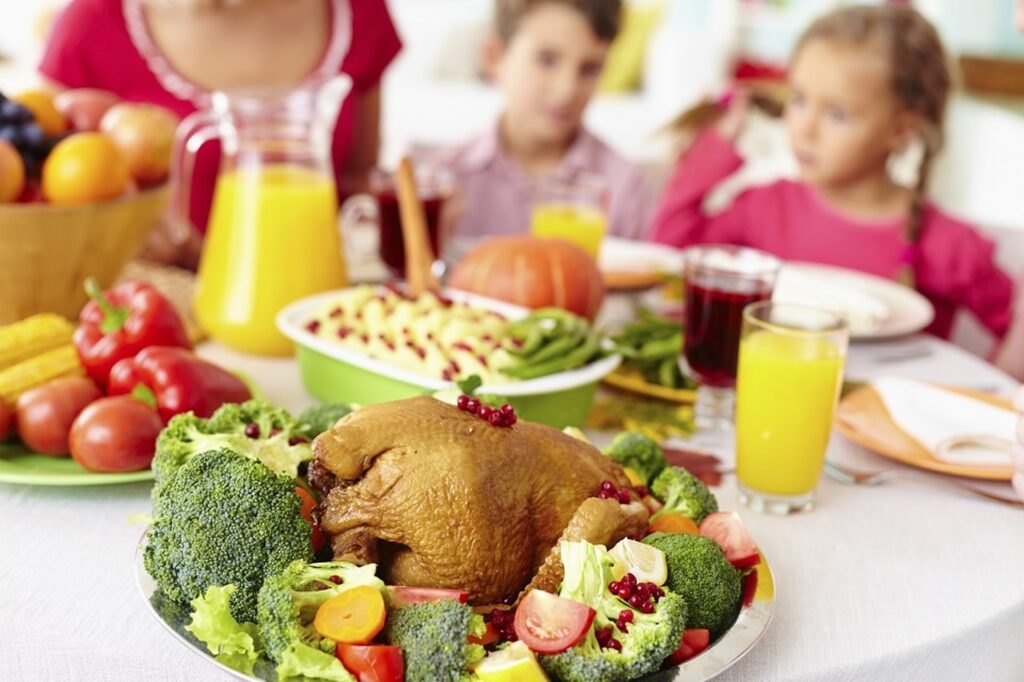 crianças ao fundo com uma mesa saudável contendo um peru, brócolis e outras verduras